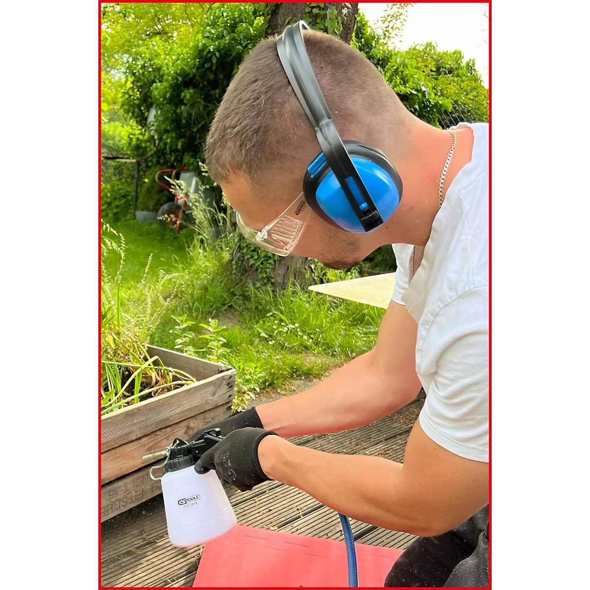 Ein Mann mit Gehörschutz und Schutzbrille hält eine Sprühflasche. Er trägt Handschuhe, im Hintergrund ist ein Garten.